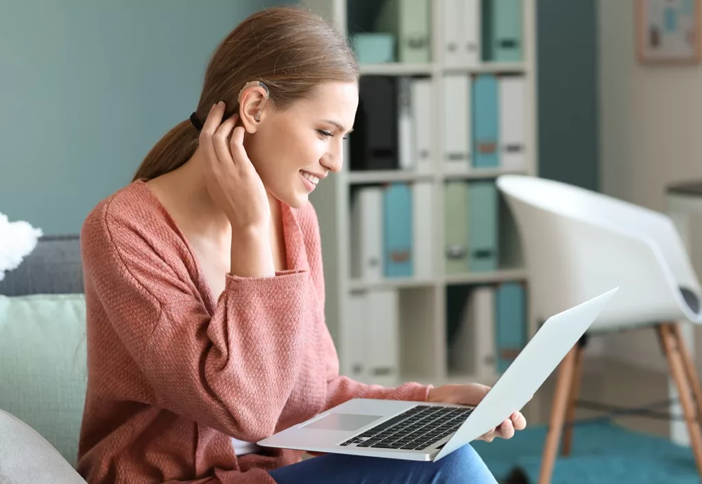 Young woman with Bluetooth hearing aids using laptop at home